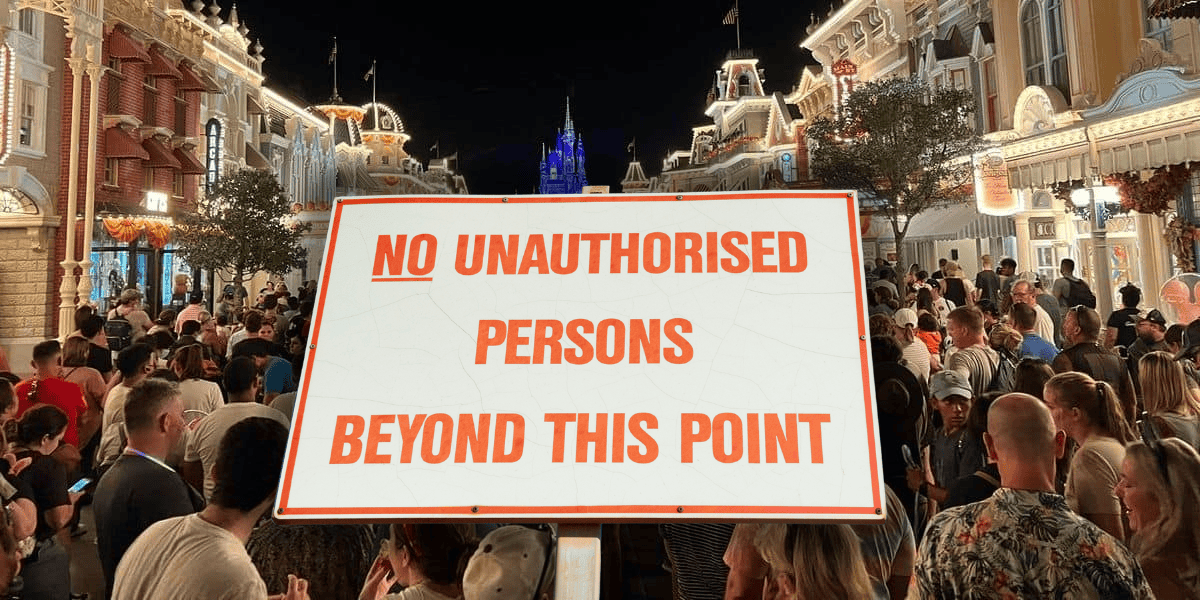 A large crowd of people stand gathered in a brightly lit area at night. A prominent sign in the foreground reads, "NO UNAUTHORISED PERSONS BEYOND THIS POINT." A Disney castle is visible in the background, illuminated and set against the night sky.