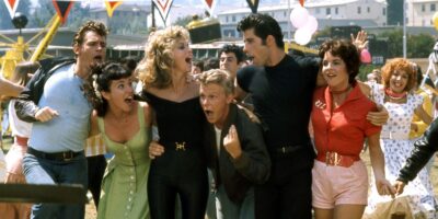 A lively group of young adults in colorful 1950s attire energetically singing and dancing at a bustling outdoor fair, full of excitement and joy.