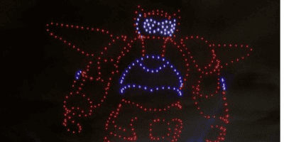 An image shows a colorful Disney drone light show creating the shape of a large robot against a dark, cloudy sky. The lights are predominantly red with some blue and white, outlining the head, body, arms, and legs of the robot.