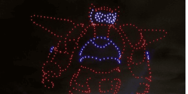 An image shows a colorful Disney drone light show creating the shape of a large robot against a dark, cloudy sky. The lights are predominantly red with some blue and white, outlining the head, body, arms, and legs of the robot.