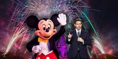 Mickey Mouse and Governor DeSantis stand in front of a castle under a spectacular fireworks display. Mickey is waving cheerfully while Governor DeSantis gestures as if presenting the event.