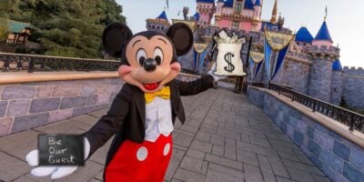 Disney's Mickey Mouse in front of a castle, holding a bag with a dollar sign and a sign reading "be our guest.