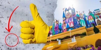 A gloved hand giving a thumbs-up is prominently displayed on the left side of the image. On the right side, there is a colorful and whimsical Disney building with various decorations and two small fountains in front, all under bright sunlight. A red arrow points to a small circled area on the left side.