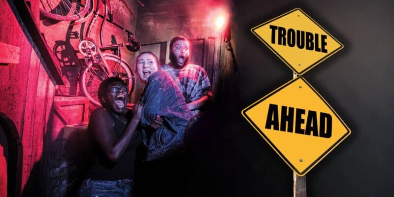 Three people with shocked expressions huddle together in a dimly lit room with red-toned lighting and old bicycles hanging on the wall. To the right, two yellow road signs stacked vertically read "TROUBLE AHEAD," hinting at a universal warning of impending challenges at Halloween Horror Nights.