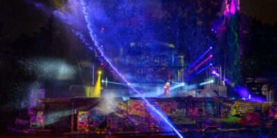 An outdoor nighttime show at Disney features colorful lights and laser effects, with mist and water sprays. A performer stands on a stage surrounded by ancient temple ruins, illuminated by vibrant beams of light against a dark background, creating a magical experience for all ages.