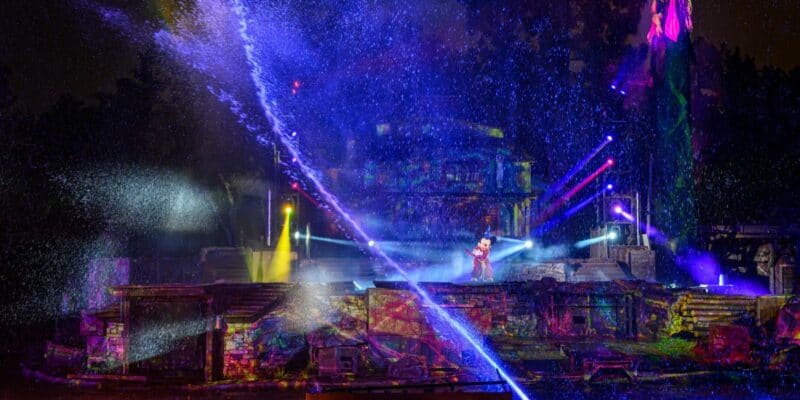 An outdoor nighttime show at Disney features colorful lights and laser effects, with mist and water sprays. A performer stands on a stage surrounded by ancient temple ruins, illuminated by vibrant beams of light against a dark background, creating a magical experience for all ages.