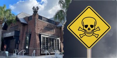A gothic-style brick building with decorative gargoyles on the roof stands under a cloudy sky, with palm trees nearby. To the right, a yellow triangular warning sign features a skull and crossbones, evoking an eerie Disney villain scene that indicates danger or poison.