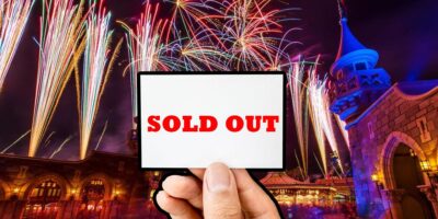 A hand holds a white sign with "SOLD OUT" written in red letters against a vibrant background of colorful fireworks illuminating the night sky above a Disney fantasy castle.