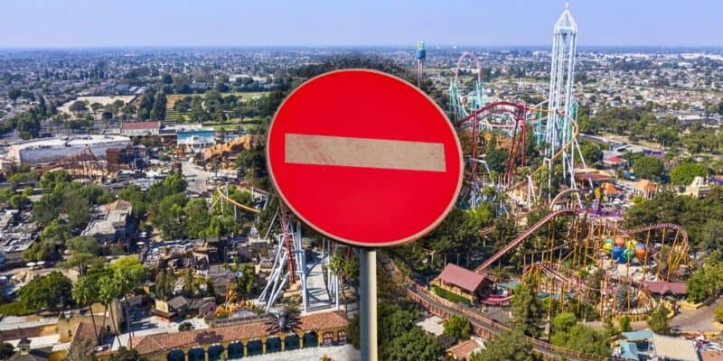 An aerial view of a theme park with various roller coasters and attractions, partially obscured by a large red and white "no entry" road sign in the foreground. The surrounding area includes trees and buildings.