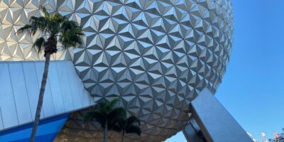 A large geodesic sphere made up of triangular panels is seen against a clear blue sky at Disney World. Two palm trees are positioned in the foreground, with the lower portion of the sphere slightly obscured by angular structures.