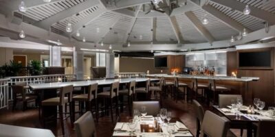 A modern, upscale restaurant at Walt Disney World with a high, white, vaulted ceiling and a circular bar area surrounded by bar stools. The dining area features neatly set tables with chairs, wine glasses, and cutlery. Soft lighting creates a cozy, inviting atmosphere.