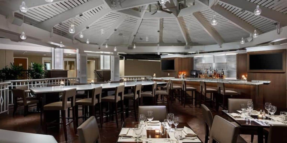 A modern, upscale restaurant at Walt Disney World with a high, white, vaulted ceiling and a circular bar area surrounded by bar stools. The dining area features neatly set tables with chairs, wine glasses, and cutlery. Soft lighting creates a cozy, inviting atmosphere.