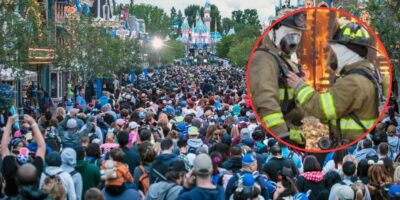 A crowded amusement park, reminiscent of Disneyland, bustling with numerous visitors wearing casual clothing and headgear. Inset within a red circle is an image of two firefighters in uniform and helmets from the Fire Department, standing close to each other with one gently touching the other's chest.
