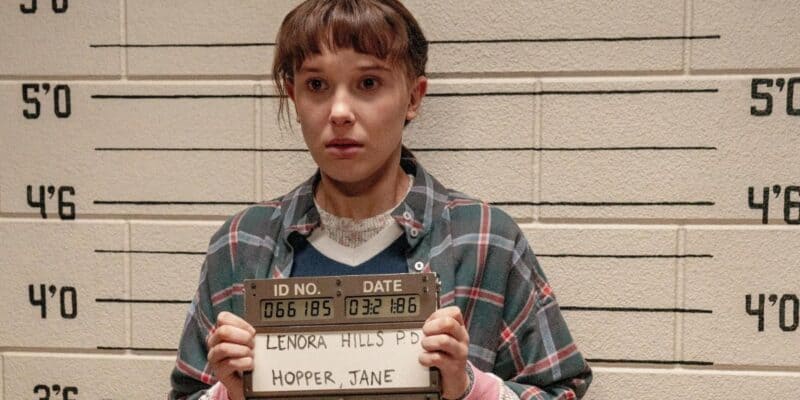 A young person with short hair, wearing a plaid shirt, stands in front of a height measurement wall holding a sign that reads "Lenora Hills P.D., Hopper, Jane." The person appears to be involved in a police booking process.