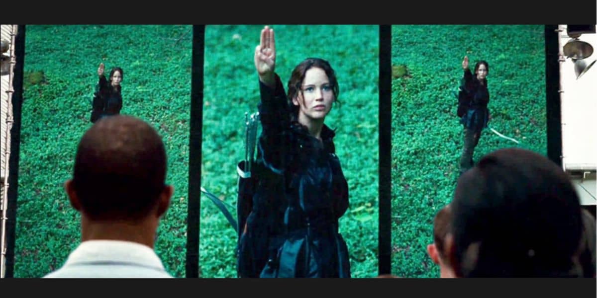 Image of multiple screens displaying a person performing a salute with three fingers raised. The screens are set against a green, forested backdrop. The scene appears to be a public broadcast. Several people are watching the screens from behind.
