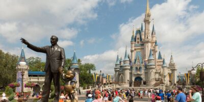 Statue of a man pointing, accompanied by a cartoon mouse, in front of a fairytale castle under a blue sky with clouds, surrounded by visitors.