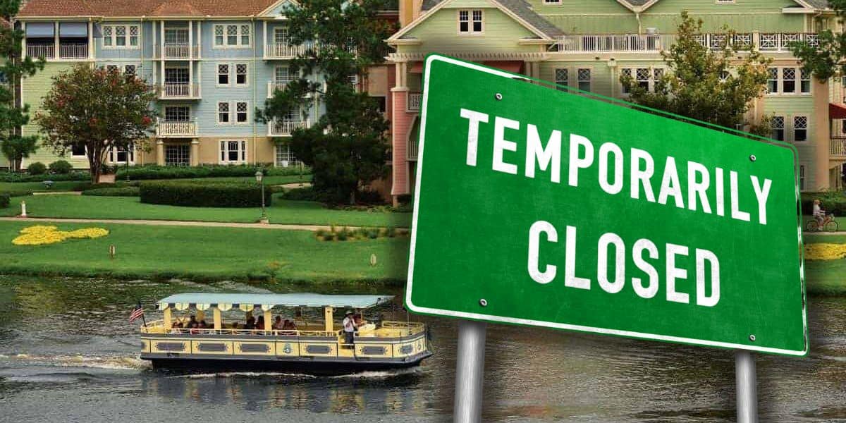 A green road sign reading "temporarily closed" in front of a scenic view featuring a colorful, Victorian-style building at Disney and a boat carrying passengers on the water.