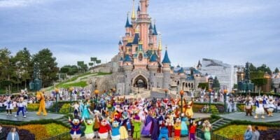 A vibrant parade at Disneyland Paris featuring numerous disney characters in colorful costumes, with a large, picturesque castle in the background and a crowd of attendees under a bright sky at this Disney Park in France.