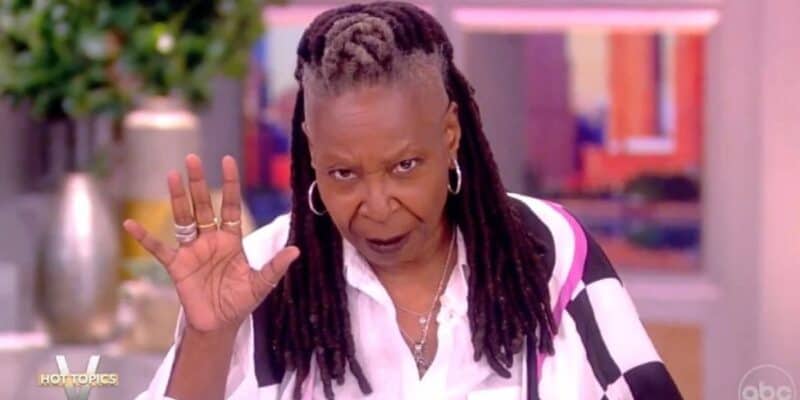 Whoopi Goldberg gestures with her hand while speaking on the set of "The View," wearing a black and white striped shirt, her hair styled in dreadlocks.