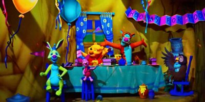 Colorful scene from Winnie the Pooh featuring various whimsical creatures at a party with balloons, a banner, and a festive table set up in a dimly lit room.