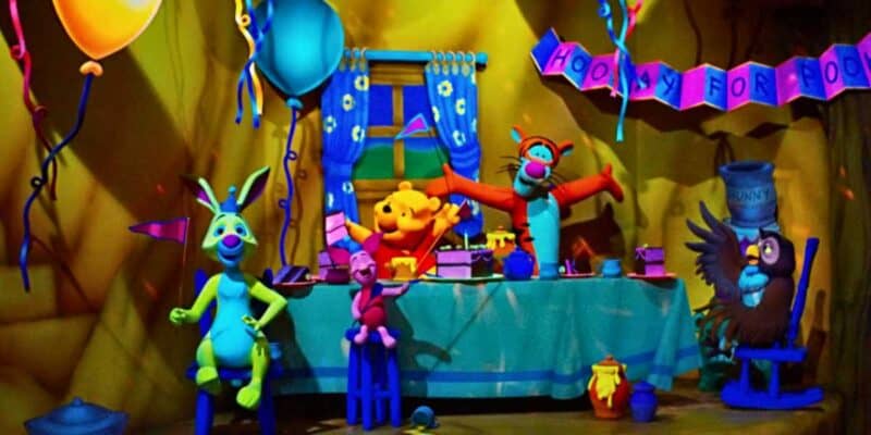 Colorful scene from Winnie the Pooh featuring various whimsical creatures at a party with balloons, a banner, and a festive table set up in a dimly lit room.
