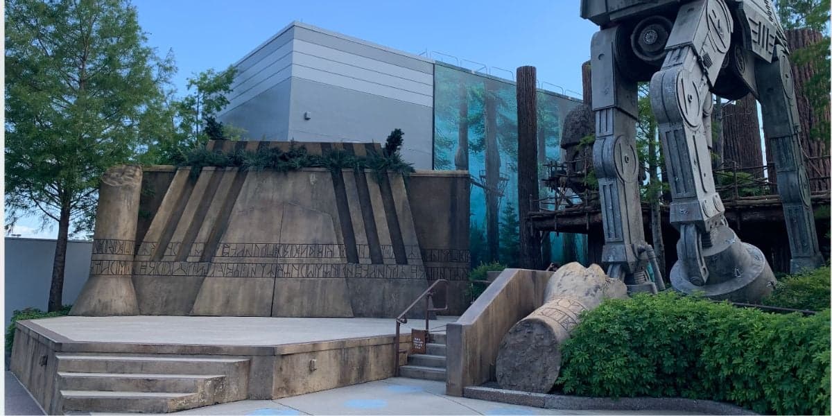 A stage flanked by themed elements including a towering robot-like structure on the right and lush green foliage, set in an outdoor park at Disney World with clear skies.