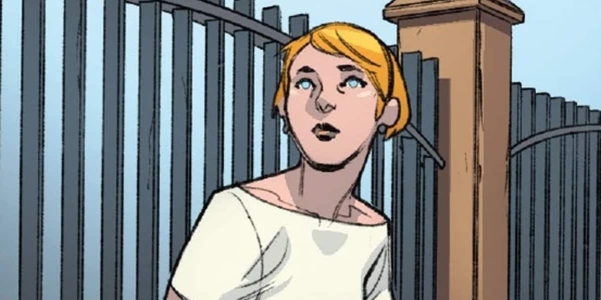 Alicia Masters with short blond hair is looking up while standing in front of a tall metal fence and a brick pillar. They are wearing a white off-the-shoulder top. The background appears to be a slightly cloudy sky.