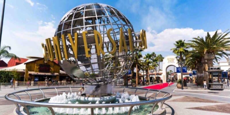 Iconic Universal Studios globe with golden "Universal" lettering in front of a fountain, under a clear sky with people displaying bad guest behavior and palm trees around, at the park entrance.