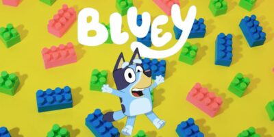 The image features the animated character Bluey, a blue heeler puppy, excitedly jumping with its arms raised against a yellow background scattered with colorful toy building blocks. The show's title, "Bluey," is prominently displayed at the top, capturing Bluey's enthusiasm for every developmental phase like a fun Lego project.