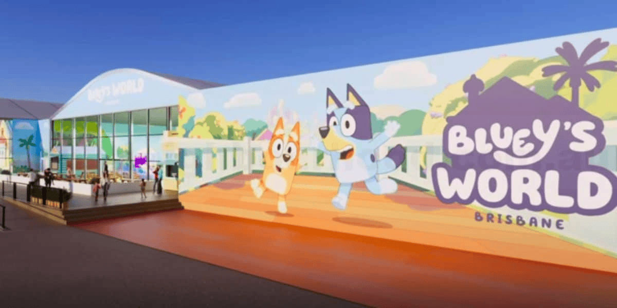 A large building with a bright mural featuring cartoon dogs Bluey and Bingo running, with the text "Bluey's World Brisbane" on it. The entrance to the building has glass doors and windows, and some people are visible near the doorway. Palm trees are in the background.