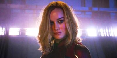 Captain Marvel (Brie Larson) with shoulder-length blonde hair and wearing a dark suit with red and gold details stands in dramatic lighting. The background features a metallic setting with bright horizontal lights, giving a futuristic ambiance, reminiscent of an MCU film scene.
