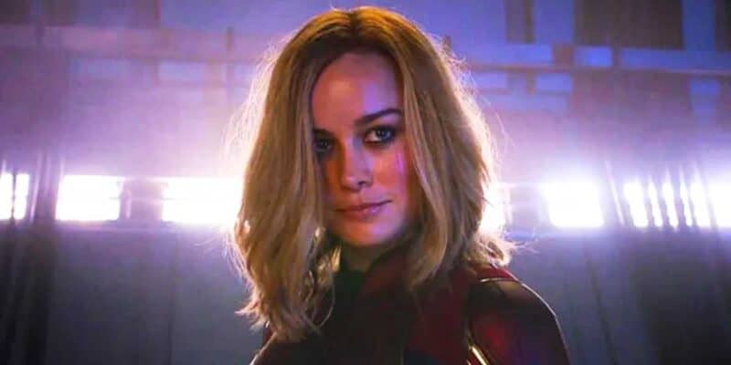 Captain Marvel (Brie Larson) with shoulder-length blonde hair and wearing a dark suit with red and gold details stands in dramatic lighting. The background features a metallic setting with bright horizontal lights, giving a futuristic ambiance, reminiscent of an MCU film scene.