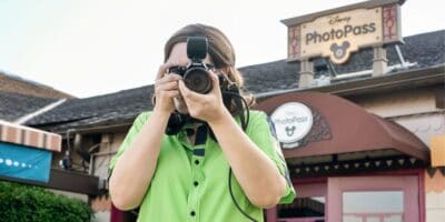 A person wearing a green shirt is holding a camera with a flash in front of their face, taking a picture. Behind them is a Disney PhotoPass sign on a building. The scene appears to be taken at a Disney theme park.
