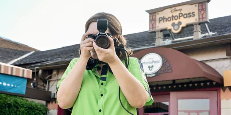 A person wearing a green shirt is holding a camera with a flash in front of their face, taking a picture. Behind them is a Disney PhotoPass sign on a building. The scene appears to be taken at a Disney theme park.