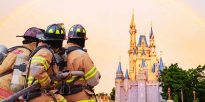 Firefighters in gear observing a scene, with a whimsical castle and a rainbow in the background, suggesting a Disney World location.