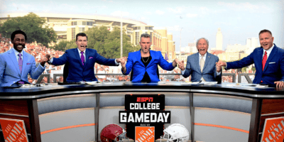 A group of five men, dressed smartly in suits with some wearing vivid colors, holds hands and smiles behind a desk with an "ESPN College GameDay" sign. The background shows a cheering stadium crowd and large buildings, indicating a lively NCAA 25 football game atmosphere. college gameday espn ncaa 25