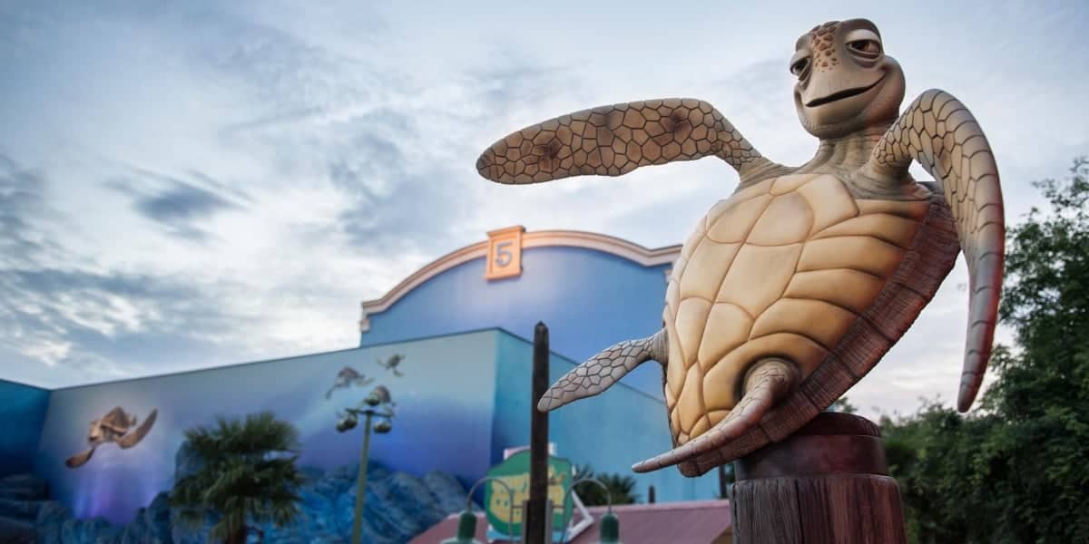 A large statue of a smiling sea turtle in the foreground, with a blue building and painted mural of flying birds in the background, under a cloudy sky, near the entrance to a fan-favorite ride.