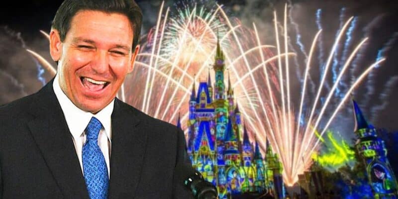 A man in a suit and tie is smiling broadly in front of a colorful night scene featuring a lit-up castle and fireworks in the sky. The castle has intricate lighting and projections, creating a celebratory atmosphere.