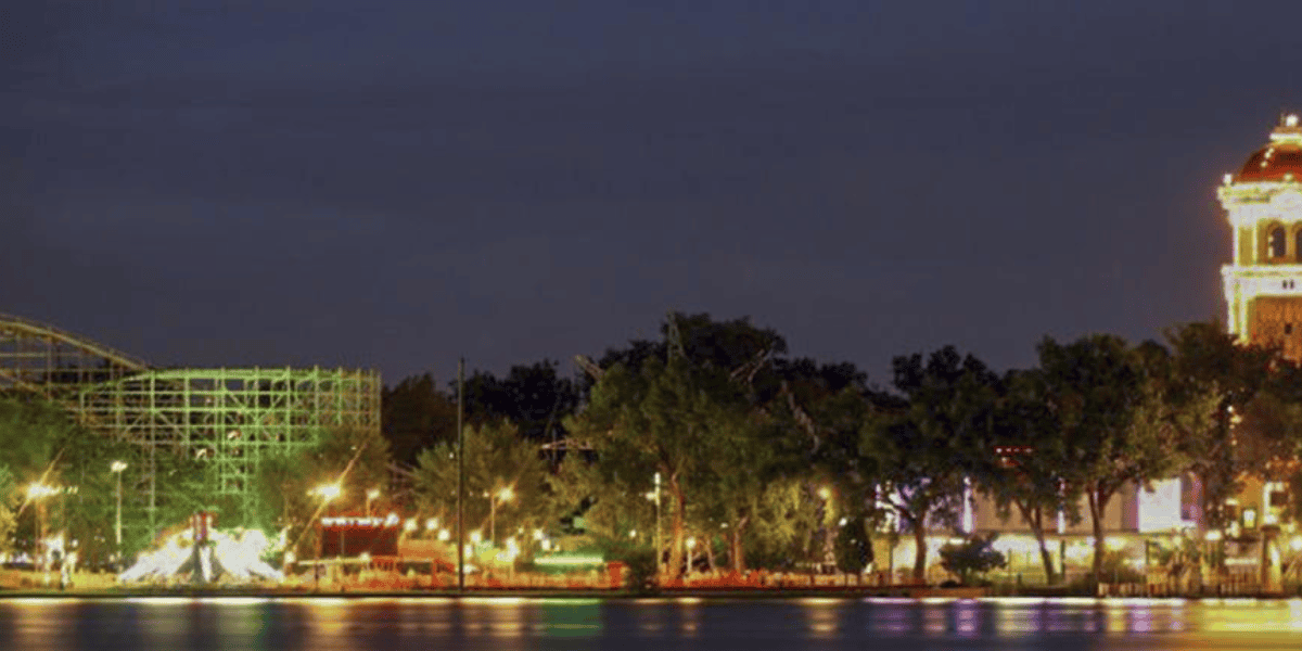 Night view of a cityscape featuring the sparkling new Disney Park Colorado with a lit Ferris wheel and roller coaster, trees, and reflections on water, under a dusk sky.