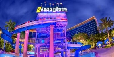 Nighttime view of Disney Resort's futuristic building with vibrant neon signs and a curving waterslide, set against a dark, starry sky with dramatic lighting.