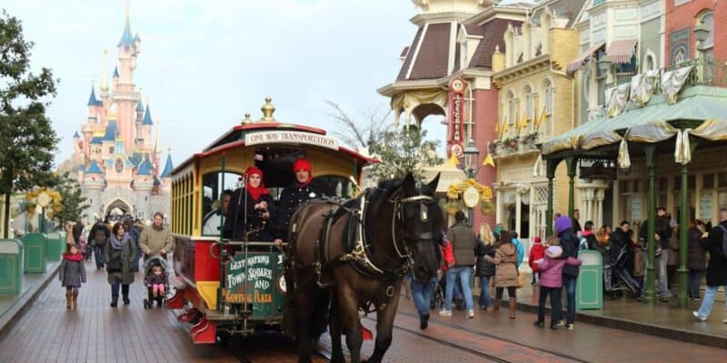 A horse-drawn trolley travels down a bustling street in a theme park with a castle in the background. The street is lined with colorful buildings, and people are walking and enjoying the scenery. Two people are riding on the trolley, which has festive decorations.