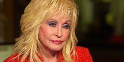 A close-up of Dolly Parton with voluminous blonde hair and striking makeup, featuring deep red lipstick and bold eye shadow. She wears a bright red outfit, exuding a warm smile.