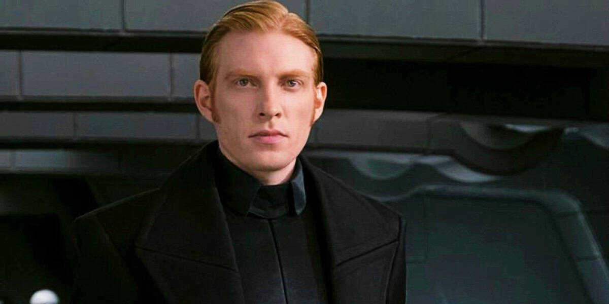 Domhnall Gleeson with light skin and straight red hair, wearing a dark high-collared jacket, stands in a room with a futuristic metallic background.