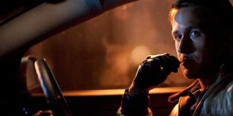 Ryan Gosling sits in a dimly lit car at night, looking towards the camera. He has a contemplative expression and is wearing leather gloves. The car's interior is dark, and there is a warm light source outside the vehicle, casting a subtle orange hue.