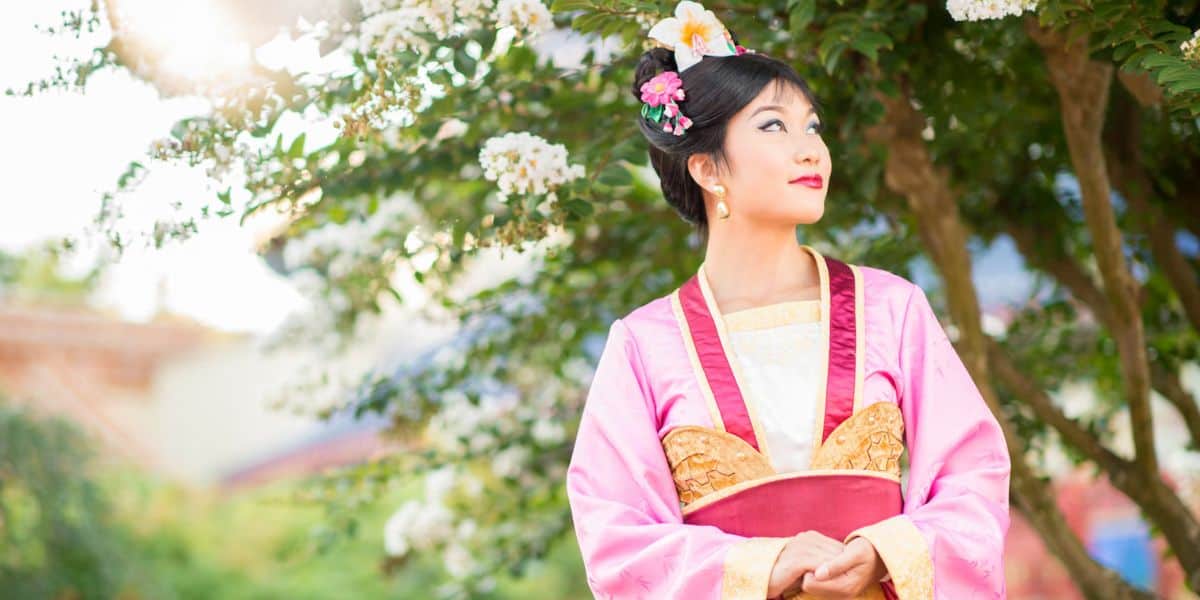 A woman in traditional korean hanbok dress, adorned with a floral hairpiece, gazes thoughtfully to the side under a tree with white flowers.