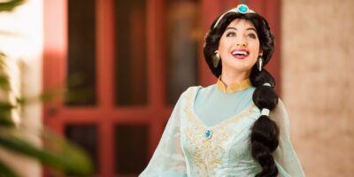 A woman dressed as princess jasmine from disney's aladdin, wearing a teal costume with gold accents and a large jewel, smiles broadly in a sunny courtyard setting.