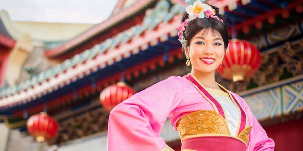 A woman in a traditional pink asian dress with floral hair accessories, smiling in front of a colorful temple decorated with red lanterns.