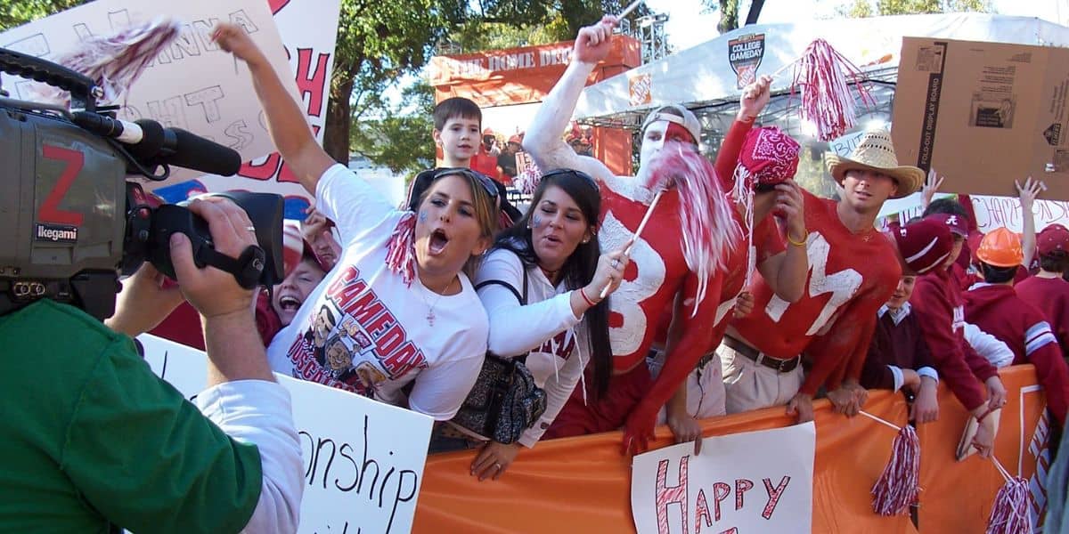 A group of enthusiastic sports fans, dressed in team colors and face paint, cheer and wave pom-poms while being filmed by a cameraman. They hold up signs and are behind a barricade at an outdoor college football event, possibly a game or rally.