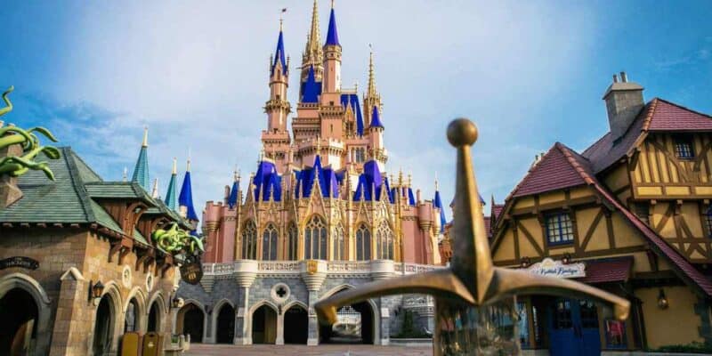 A majestic castle with blue spires is centered in the image, surrounded by charming, colorful buildings. In the foreground, a sword is embedded in a stone, reminiscent of a legendary tale. The clear and bright sky adds to the fairytale-like atmosphere that's beloved by both Disney World visitors and Florida locals.