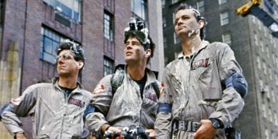 Three men in matching grey jumpsuits and specialized headgear gaze intently upwards, standing in front of a city building. Their gear and clothing are marked with signs of recent activity.